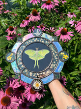 Load image into Gallery viewer, Full view in artists hand with purple coneflower backdrop . Gouache on paper, and flora set between glass with iridescent glass border in cool tones and bevels. Medallion shape with three rungs - center is painted green Luna moth on black background (circle shape), second ring is segmented circle of pressed gypsophila in clear glass, third and outer ring consists of alternating blue iridescent glass, circles with pressed flowers, and diamond bevels.