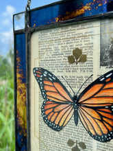 Load image into Gallery viewer, Top left angle. Orange monarch butterfly painted in Gouache on vintage floral encyclopedia, and pressed leaves set between glass with blue and gold mirrored glass border and clear beveled corners. Rectangle shape with brass hanging chain at top.