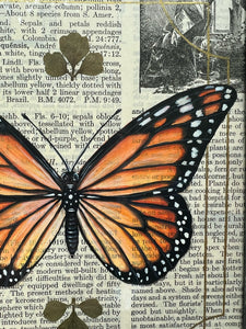 Close up of painted monarch butterfly on vintage paper with  small leaf inclusions above and below painted area. Orange monarch butterfly painted in Gouache on vintage floral encyclopedia, and pressed leaves set between glass with blue and gold mirrored glass border and clear beveled corners.  Rectangle shape with brass hanging chain at top. 