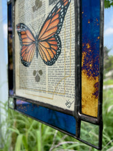 Load image into Gallery viewer, Bottom right angled photo - Orange monarch butterfly painted in Gouache on vintage floral encyclopedia, and pressed leaves set between glass with blue and gold mirrored glass border and clear beveled corners. Rectangle shape with brass hanging chain at top.