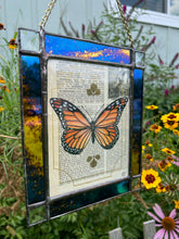 Load image into Gallery viewer, Alternate angle - Orange monarch butterfly painted in Gouache on vintage floral encyclopedia, and pressed leaves set between glass with blue and gold mirrored glass border and clear beveled corners. Rectangle shape with brass hanging chain at top.