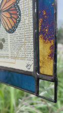 Load and play video in Gallery viewer, Video - Orange monarch butterfly painted in Gouache on vintage floral encyclopedia, and pressed leaves set between glass with blue and gold mirrored glass border and clear beveled corners.  Rectangle shape with brass hanging chain at top. 
