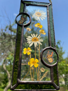 Close up - Wild grasses and daisies encased in glass with dark green glass borders segmented by two gouache monarch paintings  encased in clear glass circles. Rectangle shape with brass hanging chain at top. Center glass is clear.