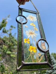 Close up - Wild grasses and daisies encased in glass with dark green glass borders segmented by two gouache monarch paintings  encased in clear glass circles. Rectangle shape with brass hanging chain at top. Center glass is clear.