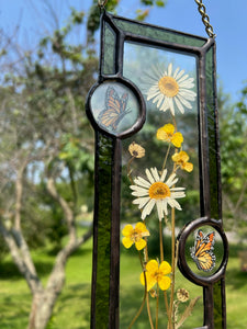 Close up - Wild grasses and daisies encased in glass with dark green glass borders segmented by two gouache monarch paintings  encased in clear glass circles. Rectangle shape with brass hanging chain at top. Center glass is clear.