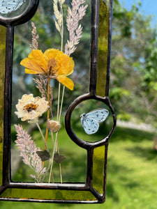 Close up - Wildflowers and yellow poppy encased in glass with yellow glass borders segmented by two light blue moth circles. Rectangle shape with brass hanging chain at top. Center glass is clear.