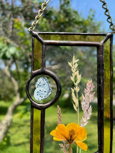 Close up - Wildflowers and yellow poppy encased in glass with yellow glass borders segmented by two light blue moth circles. Rectangle shape with brass hanging chain at top. Center glass is clear.