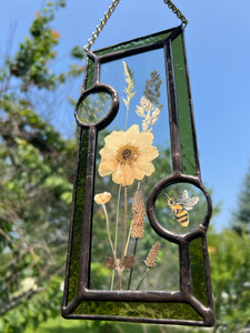 Close up - Wild grasses and Japanese anemone encased in glass with dark green glass borders segmented by one gouache honeybee painting and one golden vinyl honeycomb pattern encased in clear glass circles. Rectangle shape with brass hanging chain at top. Center glass is clear.