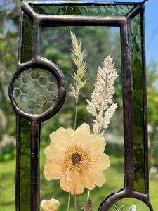 Close up - Wild grasses and Japanese anemone encased in glass with dark green glass borders segmented by one gouache honeybee painting and one golden vinyl honeycomb pattern encased in clear glass circles. Rectangle shape with brass hanging chain at top. Center glass is clear.