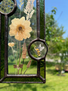 Close up - Wild grasses and Japanese anemone encased in glass with dark green glass borders segmented by one gouache honeybee painting and one golden vinyl honeycomb pattern encased in clear glass circles. Rectangle shape with brass hanging chain at top. Center glass is clear.