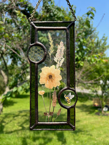 Wild grasses and Japanese anemone encased in glass with dark green glass borders segmented by one gouache honeybee painting and one golden vinyl honeycomb pattern encased in clear glass circles. Rectangle shape with brass hanging chain at top. Center glass is clear.