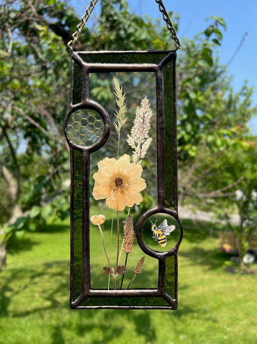 Wild grasses and Japanese anemone encased in glass with dark green glass borders segmented by one gouache honeybee painting and one golden vinyl honeycomb pattern encased in clear glass circles. Rectangle shape with brass hanging chain at top. Center glass is clear.