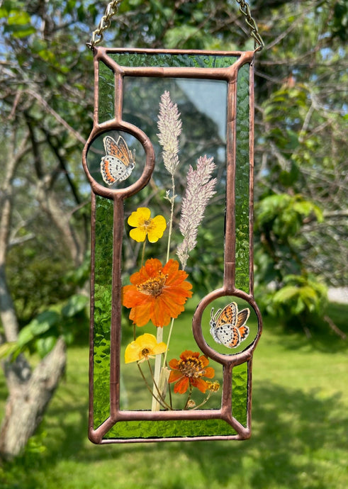 Wildflowers and orange cosmos  encased in glass with leaf green glass borders segmented by two bronze copper butterfly paintings encased in clear glass circles. Rectangle shape with brass hanging chain at top. Center glass is clear.