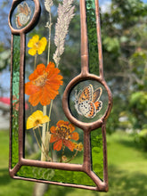 Load image into Gallery viewer, Close up - Wildflowers and orange cosmos  encased in glass with leaf green glass borders segmented by two bronze copper butterfly paintings encased in clear glass circles. Rectangle shape with brass hanging chain at top. Center glass is clear.
