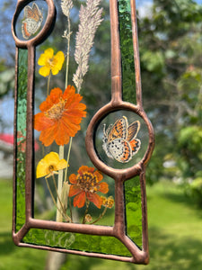 Close up - Wildflowers and orange cosmos  encased in glass with leaf green glass borders segmented by two bronze copper butterfly paintings encased in clear glass circles. Rectangle shape with brass hanging chain at top. Center glass is clear.