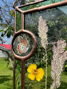 Close up - Wildflowers and orange cosmos  encased in glass with leaf green glass borders segmented by two bronze copper butterfly paintings encased in clear glass circles. Rectangle shape with brass hanging chain at top. Center glass is clear.