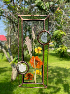 Close up - Wildflowers and orange cosmos  encased in glass with leaf green glass borders segmented by two bronze copper butterfly paintings encased in clear glass circles. Rectangle shape with brass hanging chain at top. Center glass is clear.