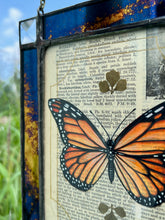 Load image into Gallery viewer, Top left angle. Orange monarch butterfly painted in Gouache on vintage floral encyclopedia, and pressed leaves set between glass with blue and gold mirrored glass border and clear beveled corners.  Rectangle shape with brass hanging chain at top. 
