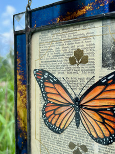 Top left angle. Orange monarch butterfly painted in Gouache on vintage floral encyclopedia, and pressed leaves set between glass with blue and gold mirrored glass border and clear beveled corners.  Rectangle shape with brass hanging chain at top. 