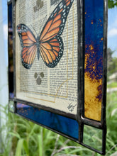 Load image into Gallery viewer, Bottom right angled photo - Orange monarch butterfly painted in Gouache on vintage floral encyclopedia, and pressed leaves set between glass with blue and gold mirrored glass border and clear beveled corners.  Rectangle shape with brass hanging chain at top. 
