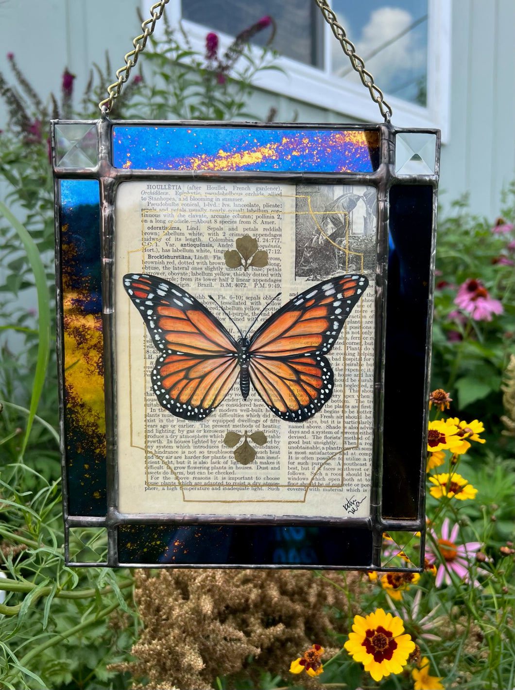 Orange monarch butterfly painted in Gouache on vintage floral encyclopedia, and pressed leaves set between glass with blue and gold mirrored glass border and clear beveled corners.  Rectangle shape with brass hanging chain at top. 