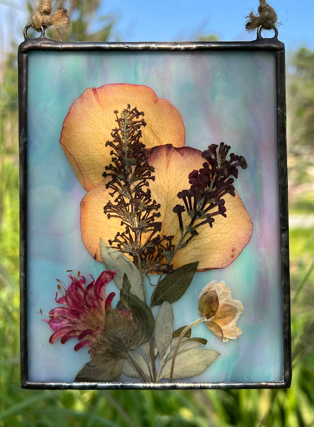 Pressed rose petals, butterfly bush, bee balm, and Japanese anemone bud with iridescent blue/pink/purple glass backing. Rectangle shape with hanging twine at top.