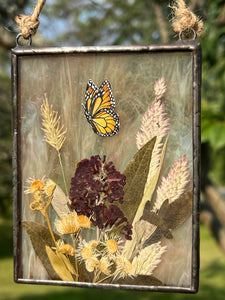 Another full view in bright indirect lighting. Painted monarch butterfly in gouache on paper with pressed butterfly bush flower, aster, and wild grasses with iridescent wispy pink glass backing. Rectangle shape with twine hanging. 