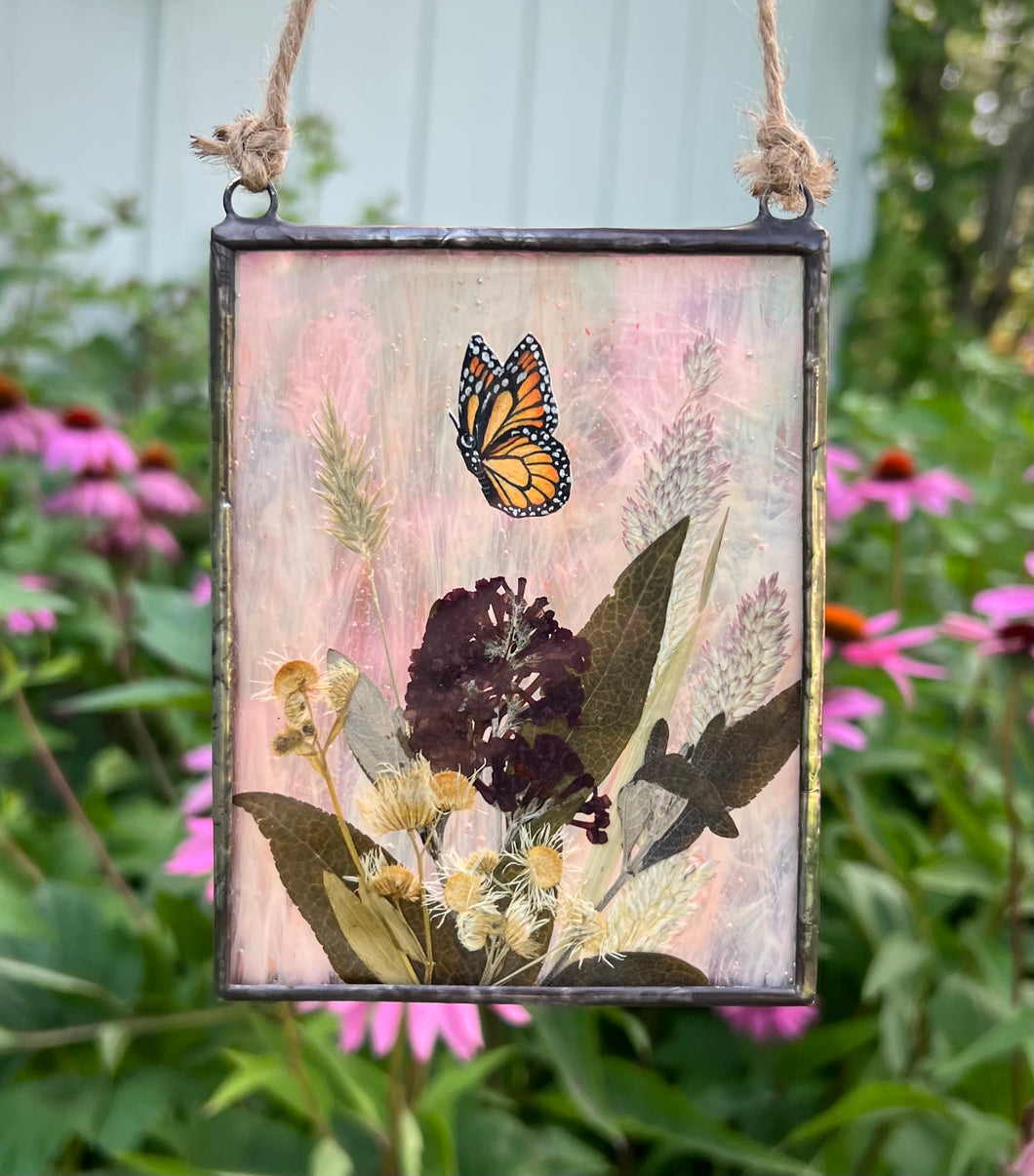 Painted monarch butterfly in gouache on paper with pressed butterfly bush flower, aster, and wild grasses with iridescent wispy pink glass backing. Rectangle shape with twine hanging.