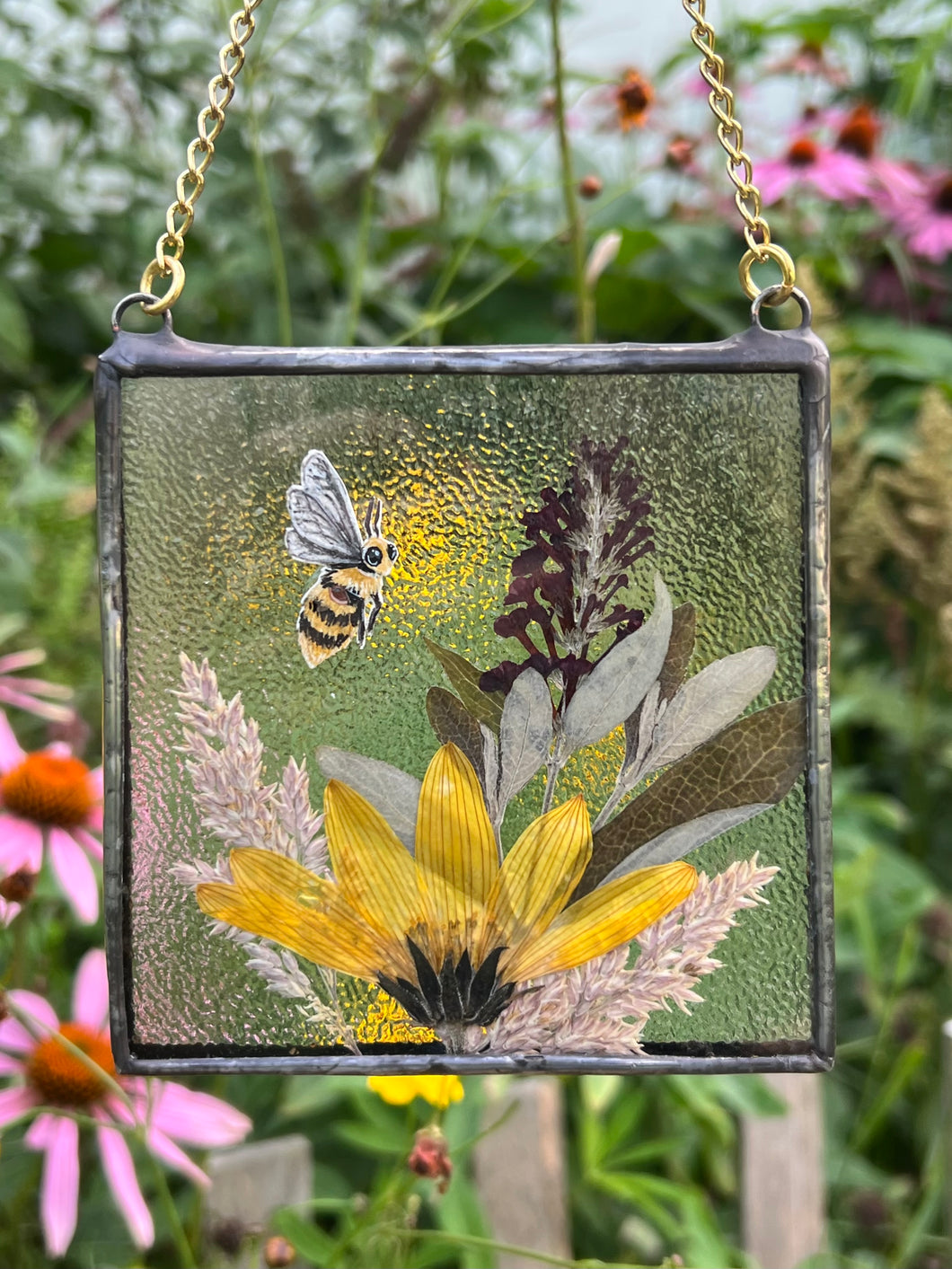 Honeybee painted in gouache on paper with wildflower grasses and  yellow sunflower and butterfly bush with clear textured glass backing. Square shape with gold chain hanger.