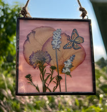 Load image into Gallery viewer, Alternate view with sun backlight to show purple glass backing. Monarch butterfly painted in gouache on paper with rose petals, lavender, and purple clover with solid wispy light purple glass backing. Square shaped artwork with twine hanging at top.
