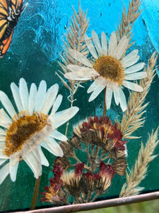 Close up view - Monarch butterfly painted in gouache on paper with pressed shasta daisies, orange hawkweed, and wild grasses with transparent blue glass backing. Square shape with gold hanging chain. Flower close up.