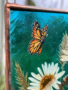 Monarch butterfly painted in gouache on paper with pressed shasta daisies, orange hawkweed, and wild grasses with transparent blue glass backing. Square shape with gold hanging chain. Close up view. 