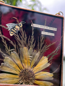 View of piece with sun as backdrop to show maroon color. Honeybee painted in gouache on paper with wildflower grasses and  yellow flower with dark purple/maroon glass backing. Artwork is square shape with chain hanging.