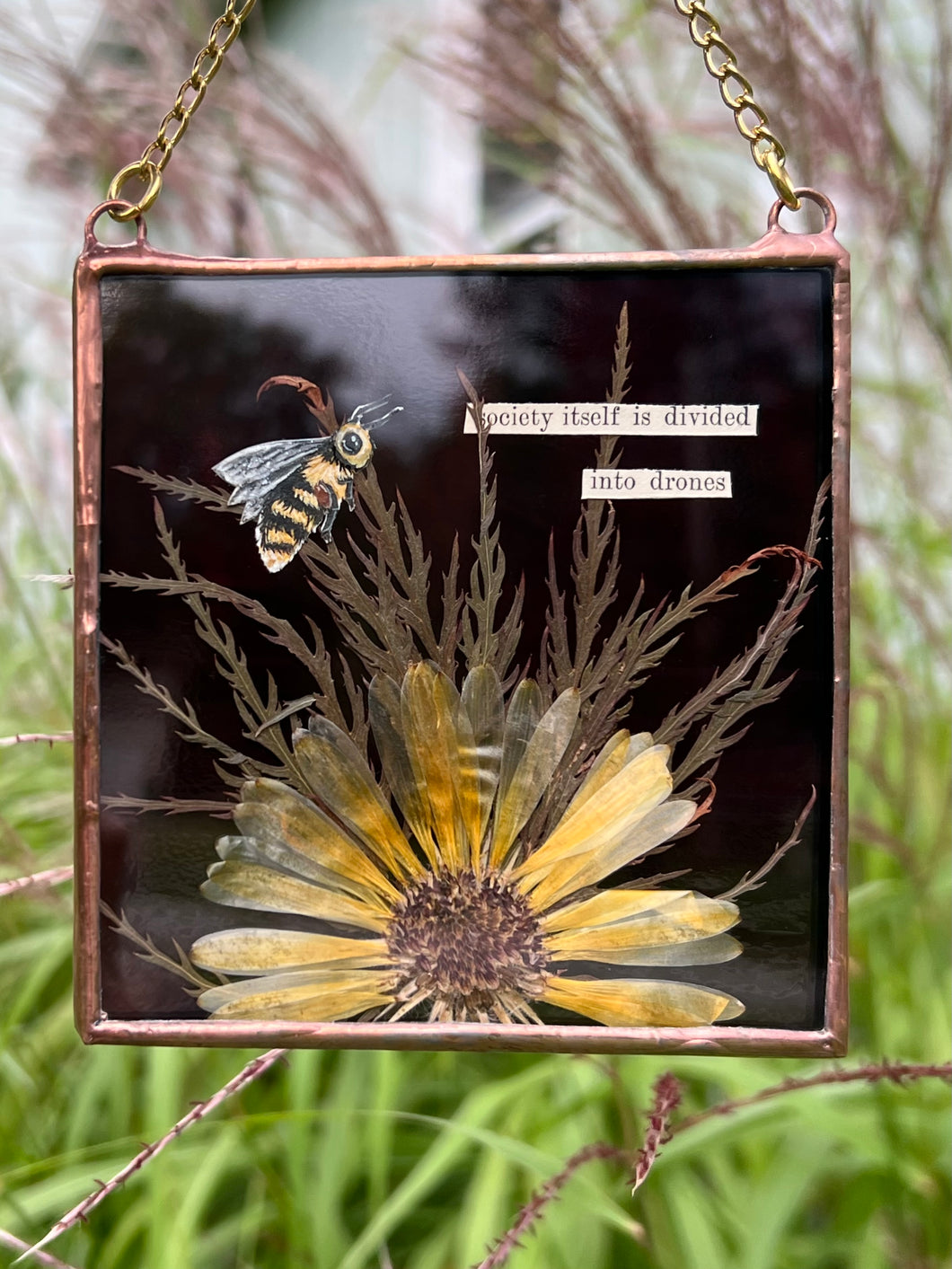 Honeybee painted in gouache on paper with wildflower grasses and  yellow flower with dark purple/maroon glass backing. Artwork is square shape with chain hanging.