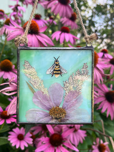 Full view with purple coneflower backdrop - Honeybee painted in gouache on paper with wildflower grasses and  purple/pink cosmos flower with blue/green glass backing. Square shape with twine hanging.
