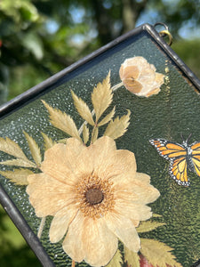Close up of Japanese anemone flower. Monarch butterfly painted in gouache on paper with pressed Japanese anemone with textured clear glass backing Square shaped artwork with gold hanging chain.
