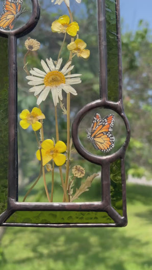 Video - Wild grasses and daisies encased in glass with dark green glass borders segmented by two gouache monarch paintings  encased in clear glass circles. Rectangle shape with brass hanging chain at top. Center glass is clear.