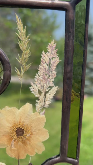 Video - Wild grasses and Japanese anemone encased in glass with dark green glass borders segmented by one gouache honeybee painting and one golden vinyl honeycomb pattern encased in clear glass circles. Rectangle shape with brass hanging chain at top. Center glass is clear.