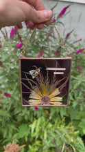 Load and play video in Gallery viewer, Video of artwork - Honeybee painted in gouache on paper with wildflower grasses and  yellow flower with dark purple/maroon glass backing. Artwork is square shape with chain hanging.
