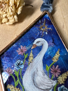 Close up of painted goose - White goose painting on blue dyed paper with pink, blue, and yellow flowers surrounding it at the base. Gold painted sun above goose. Diamond shape. Gouache painting on hand dyed paper in beveled glass.